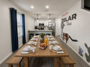 Un comedor con una larga mesa de madera dispuesta para el desayuno, una cocina al fondo y un mural en la pared con un cactus y las palabras 'TOLAR' y 'Texas'.