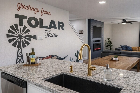 Une cuisine moderne avec un plan de travail en granit, un robinet doré et un évier. Un mur est décoré d’une fresque représentant un moulin à vent, ainsi que du texte suivant : « Salutations de TOLAR, Texas ».