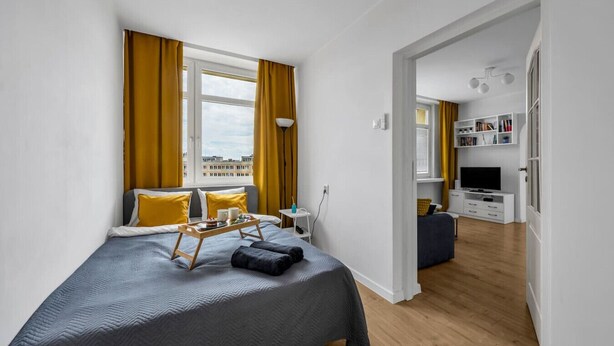 A bedroom with a bed, a bedside table, a tray with items, a window with curtains, and a view of buildings. - Downtown Apt - Top Location, Metro nearby (Warszawa)
