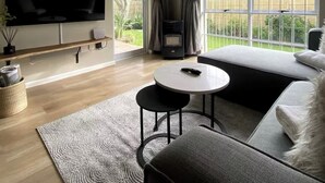Una sala moderna con un televisor de pantalla plana grande, un sofá, una mesa de centro y vistas a un jardín a través de amplias ventanas.