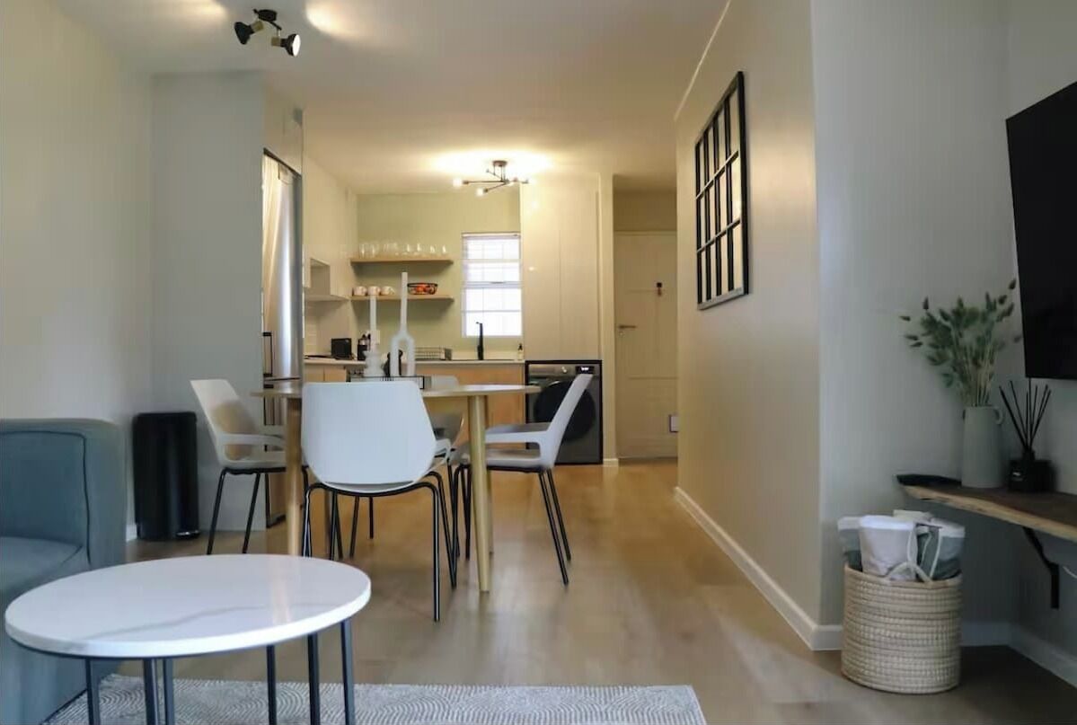Un salon moderne avec une table ronde blanche, un canapé gris et un coin repas avec des chaises, ainsi qu’une petite cuisine en arrière-plan.