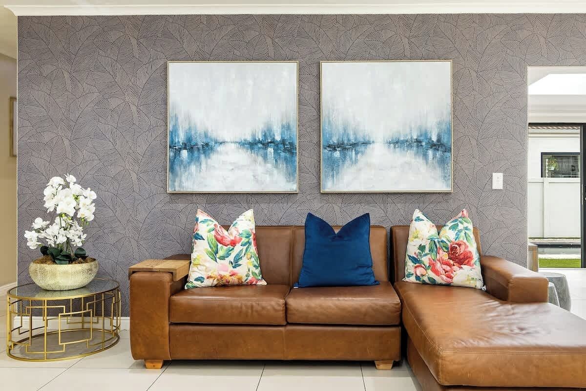 A brown leather sofa with floral and solid blue cushions, flanked by two abstract paintings on a patterned wall.
