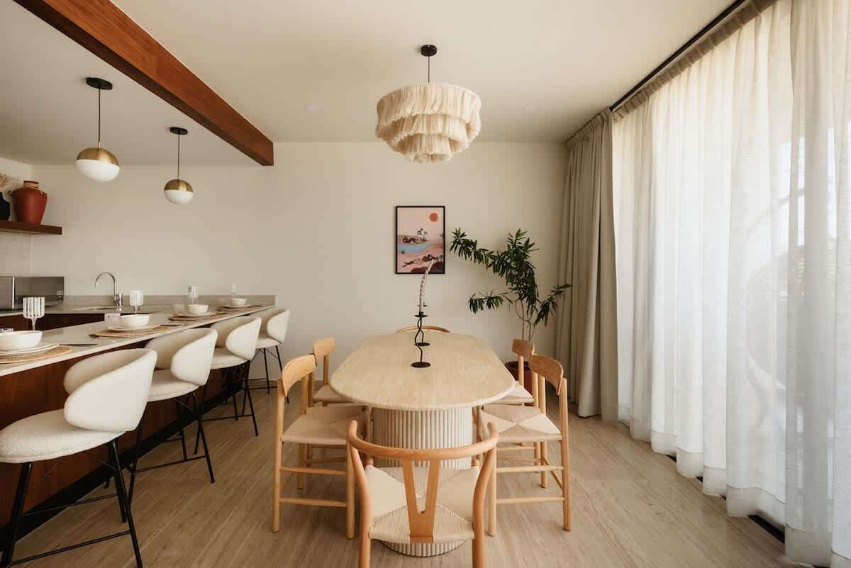 A modern dining area with a round table, white chairs, pendant lights, and a large window with sheer curtains.