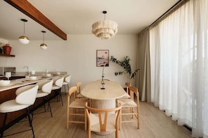A modern dining area with a round table, white chairs, pendant lights, and a large window with sheer curtains.