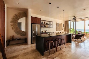 A modern kitchen with a central island, bar stools, pendant lights, and a view of the ocean.