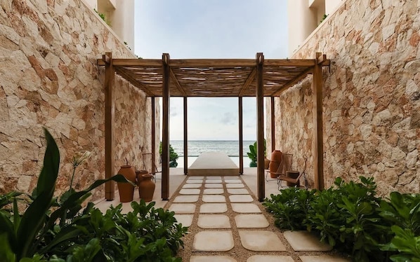 A stone pathway leading to a beach view, a wooden pergola, and potted plants.