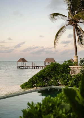 A tranquil beach scene with a pool, two thatched-roof structures, and a palm tree.