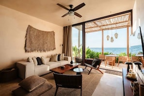 A modern living room with a sofa, coffee table, and chairs, a ceiling fan, and a large window with a view of the ocean.