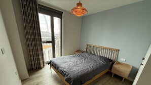 Un dormitorio con cama, piso de madera, una mesita de noche, una lámpara colgante y vista a un edificio a través de una ventana.