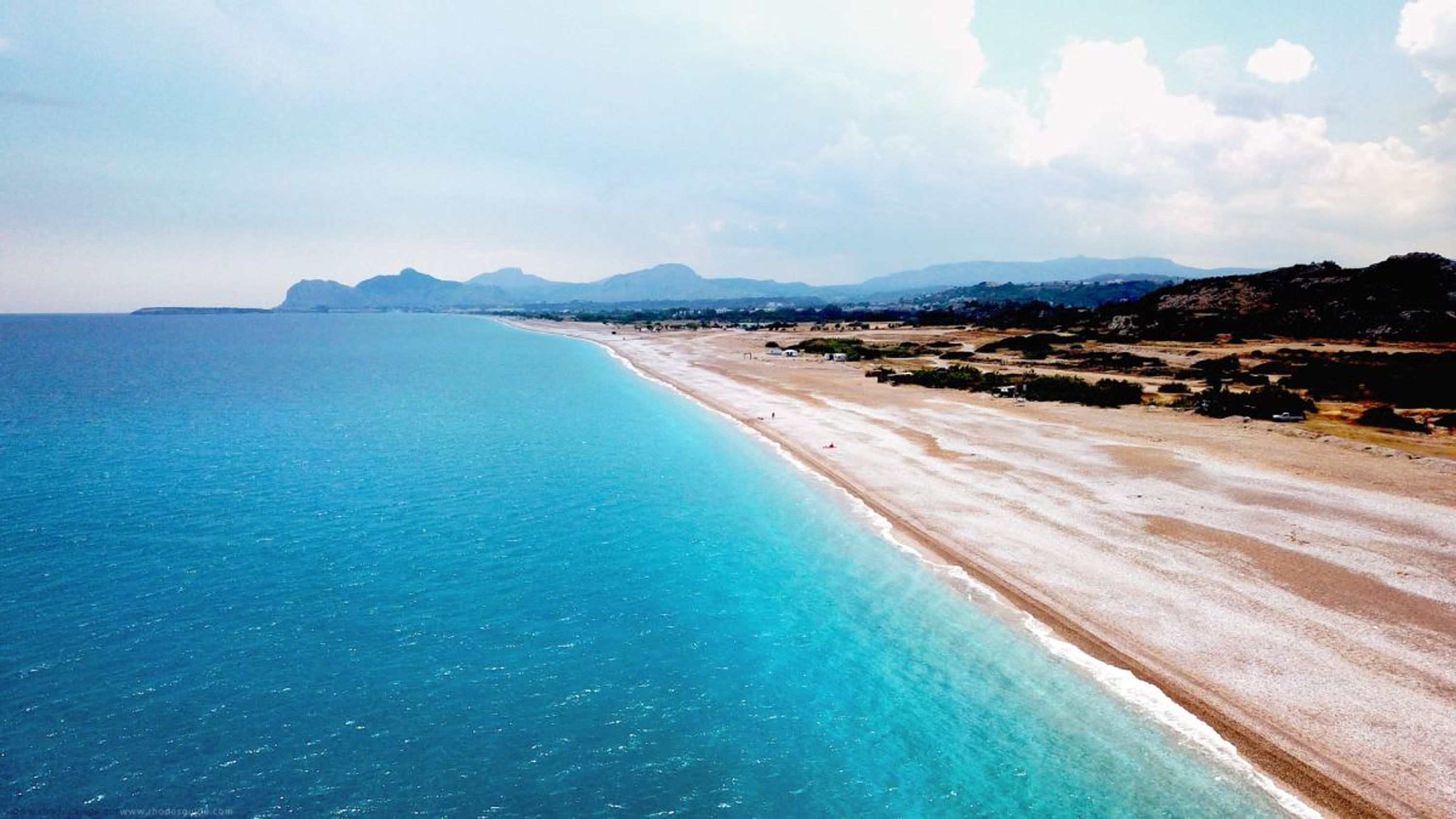 A coastal beach with a clear blue sea, sandy shore, and distant mountains.