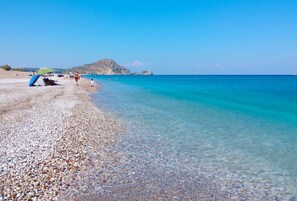 A pebbled beach with clear turquoise water, a distant rocky island, and a few people and umbrellas. - Sinani Villas Rhodes - Private Pool Retreat (Rhodes)