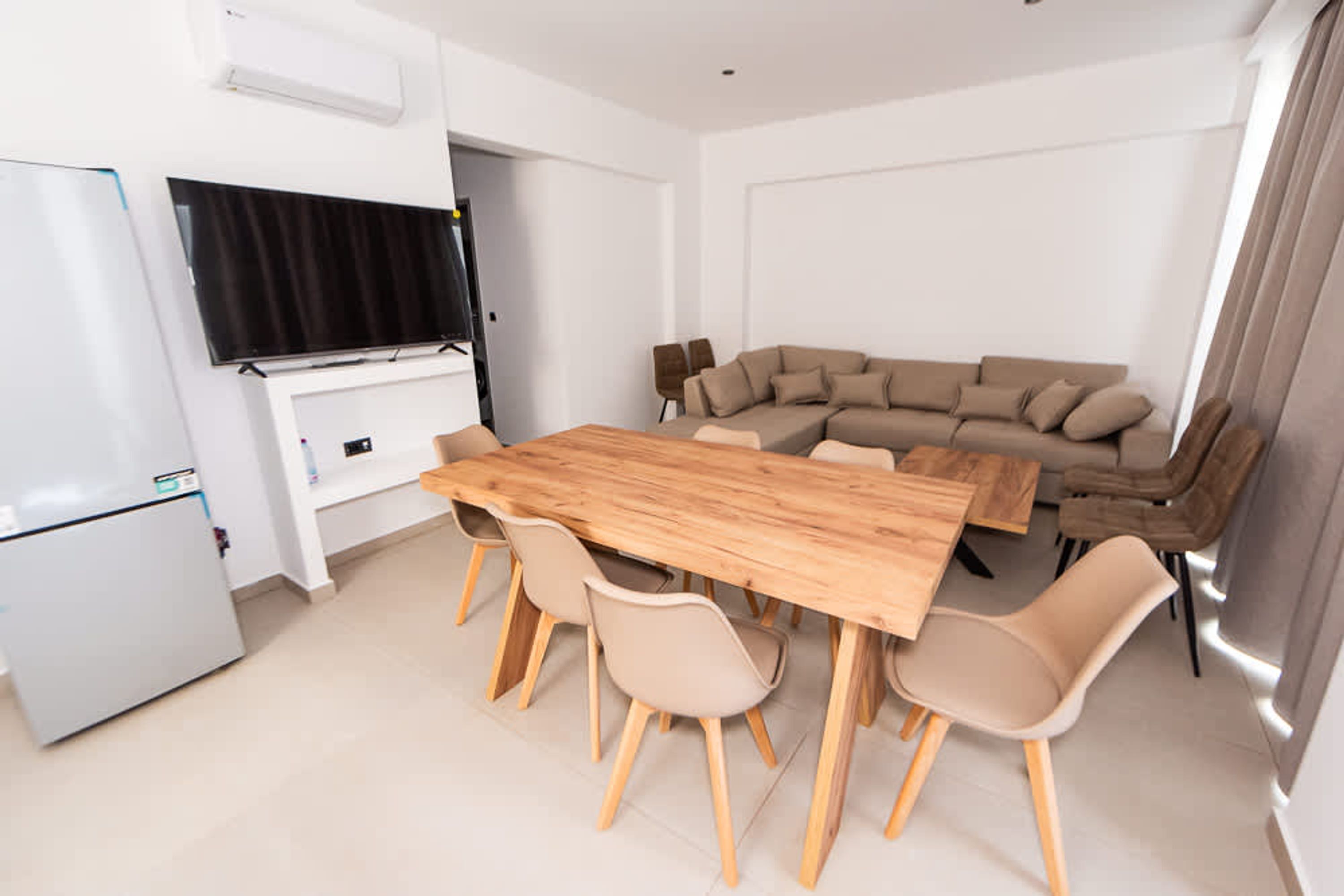 A modern living room with a wooden dining table, beige chairs, a beige sofa, and a flat-screen TV mounted on the wall.