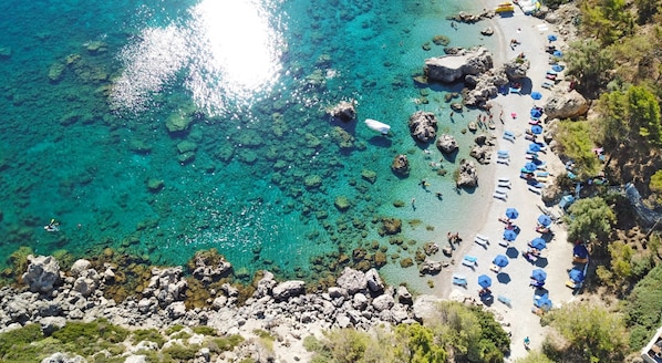 Une vue aérienne d’une plage aux eaux turquoise et limpides, parsemée de rochers et bordée d’une rangée de parasols.
