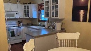Cocina compacta con armarios blancos, un microondas, un horno y un lavabo. Hay una mesa de comedor con dos sillas y un bol sobre ella.
