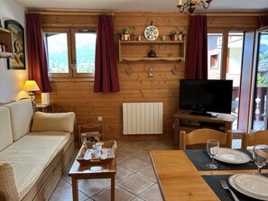 Interior acolhedor em madeira, com um sofá, uma mesa de jantar posta para dois, uma televisão e uma janela com cortinas.