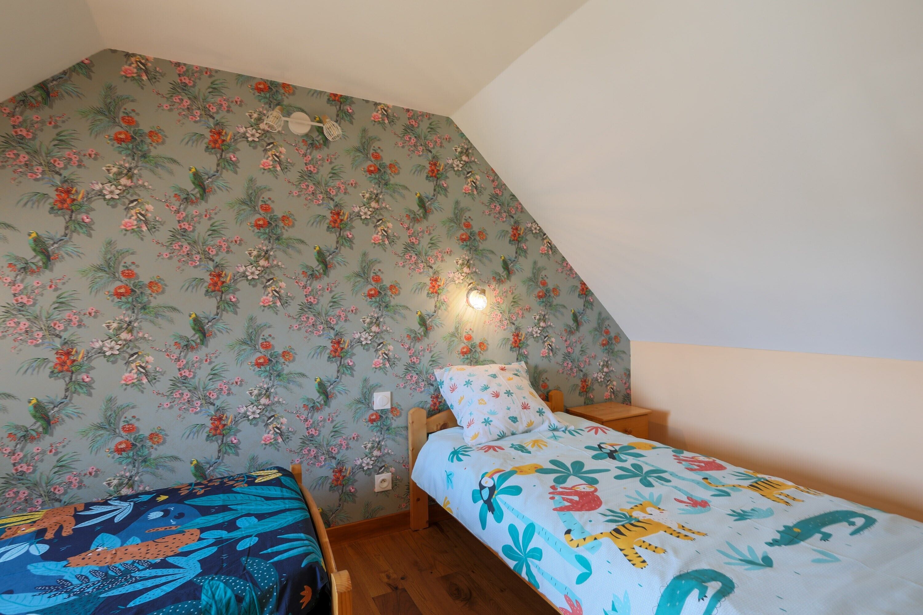 Ein Schlafzimmer mit schrägen Wänden, floralem Wandpapier, einem Bett mit bunter Bettdecke und einem Nachttisch mit Lampe.