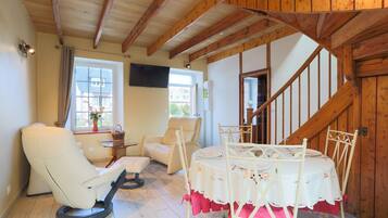 A room with a wooden ceiling, a white chair, a dining table with a floral tablecloth, and a staircase.