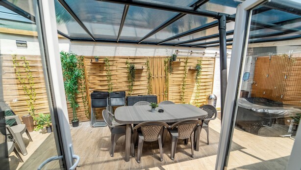 An outdoor patio with a glass enclosure, a round table with chairs, and a wooden fence with climbing plants.
