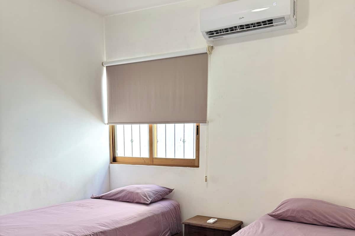 A room with two beds, a window with a blind, an air conditioning unit, and a wooden nightstand.