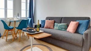 Ein modernes Wohnzimmer mit einem grauen Sofa, blauen Sesseln, einem Esstisch mit Kuchen und Champagner und Blick durch ein großes Fenster.