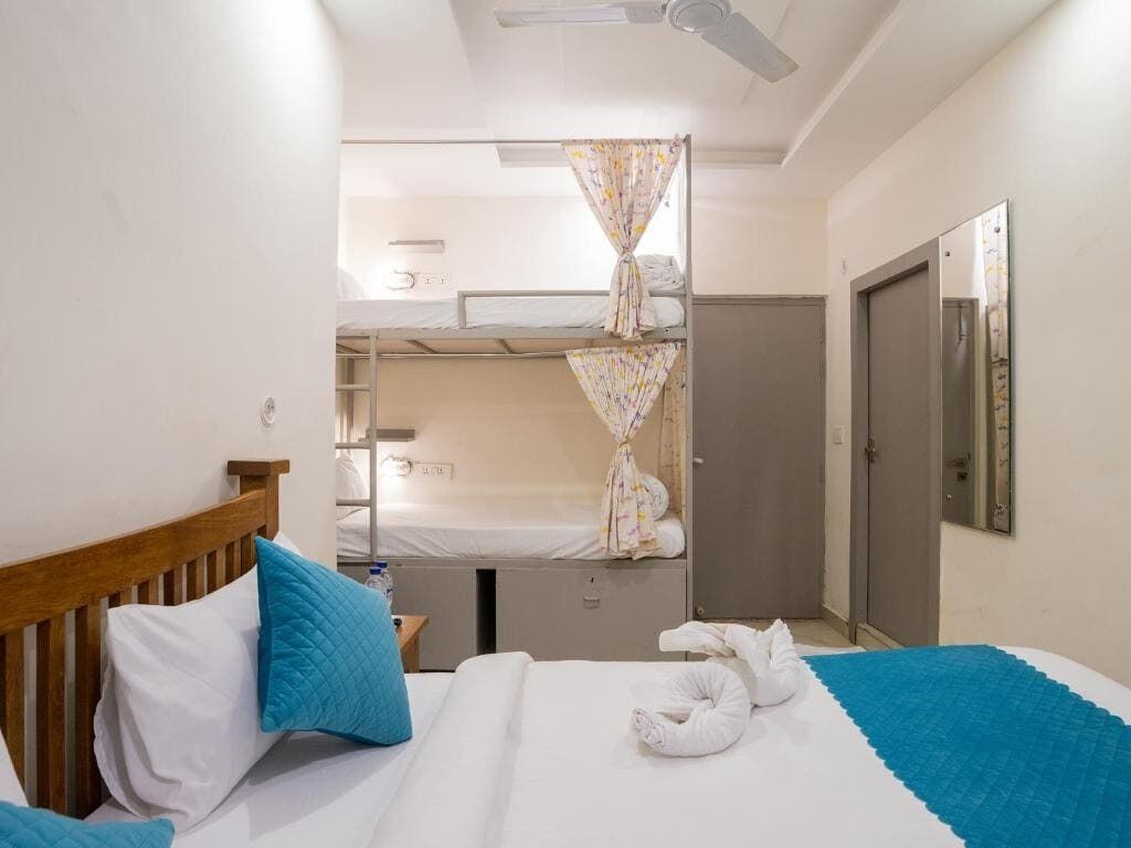 A hotel room with a single bed, a bunk bed in the background, a ceiling fan, and a door.