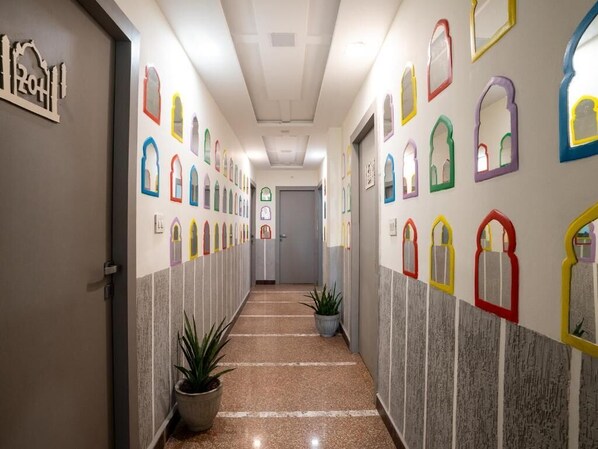 A hallway with a series of colorful, framed window designs on the walls, a potted plant, and a door at the end. - MTJ Hotels (Agra)