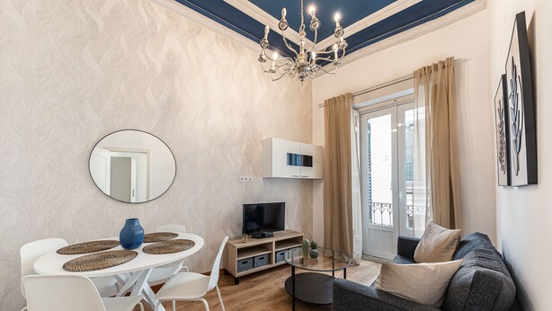 A modern living room with a dining area, a chandelier, a flat-screen TV, and a large window with curtains.