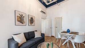 A living room with a grey sofa, a glass coffee table, and a dining area with white chairs and a blue vase.