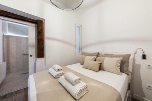 A bedroom with a bed, towels, a bedside table, a wall-mounted lamp, and a door leading to a bathroom.