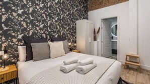 A bedroom with a black floral wallpaper, a white wardrobe, a wooden nightstand with a lamp, and a neatly made bed with rolled towels. - Waou Chamberi VIII - Short Term Rental (Madrid)