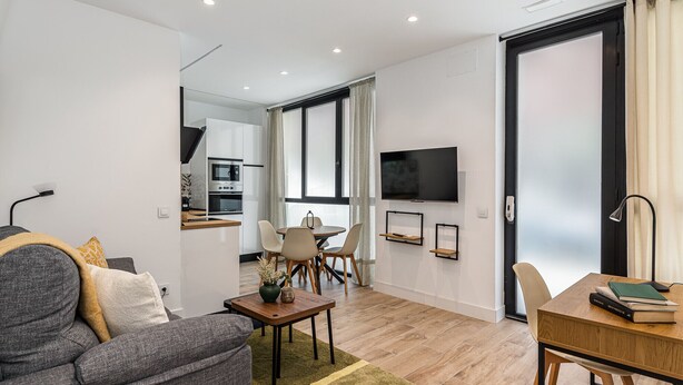 A modern living room with a sofa, coffee table, and dining area. There is a kitchen with an oven and microwave, and a TV mounted on the wall. - Waou Chamberi VIII - Short Term Rental (Madrid)
