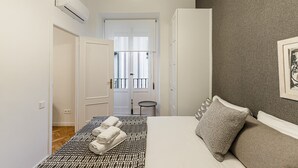 Un dormitorio con una cama, una mesita pequeña y una silla. Hay un equipo de aire acondicionado en la pared y una puerta que da a otra habitación.