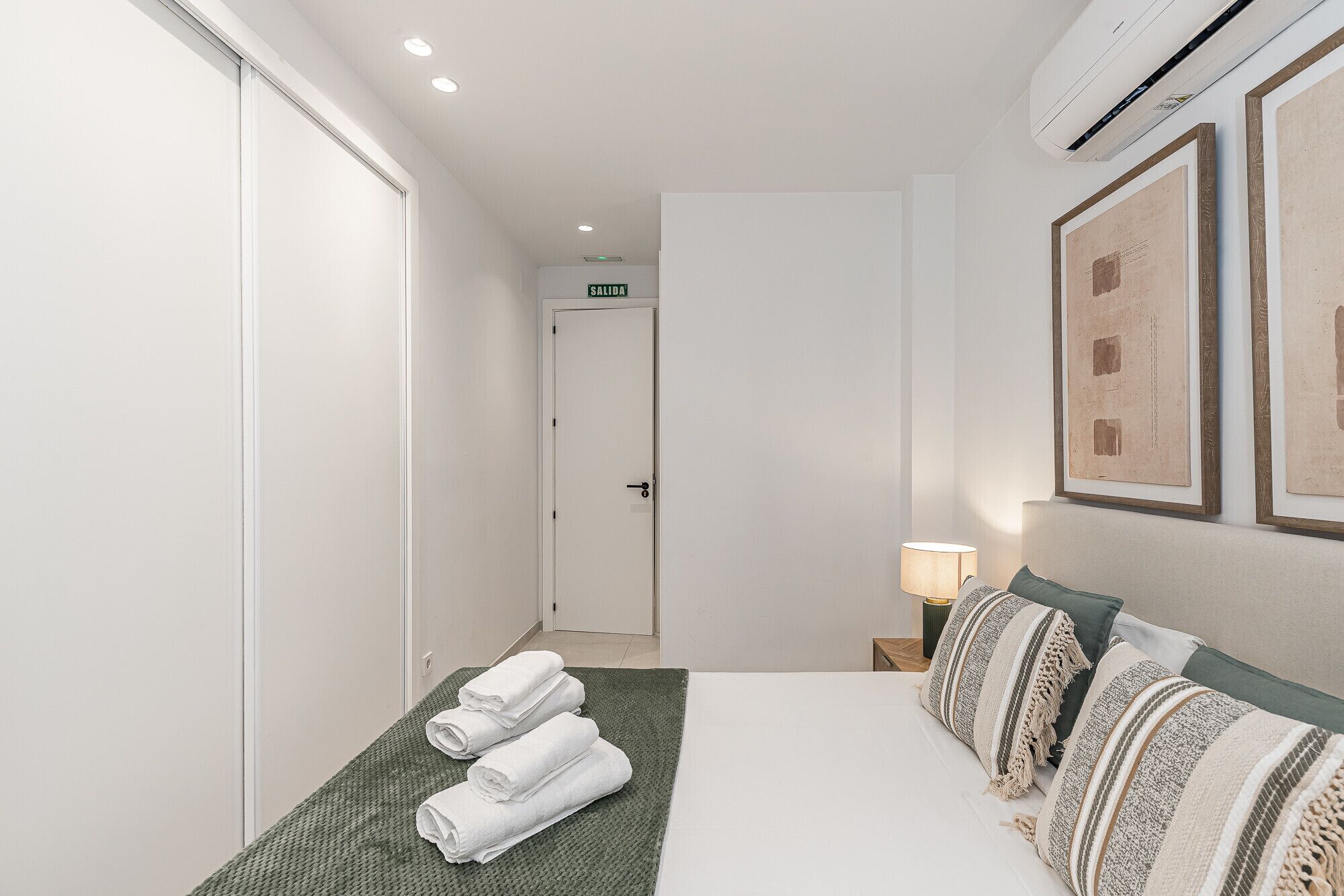 Habitación de hotel con cama, alfombra verde texturizada, dos toallas enrolladas, lámpara de noche y cuadros en la pared.