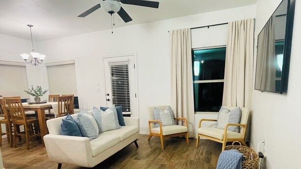 A living room with a sofa, two armchairs, a dining table with chairs, a ceiling fan, and a large window with curtains.