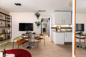 Eine moderne Küche mit weißen Schränken, einem eingebauten Ofen und einer Spüle. Es gibt einen Essbereich mit einem runden Tisch und Stühlen, sowie ein an der Wand montierter Fernseher.