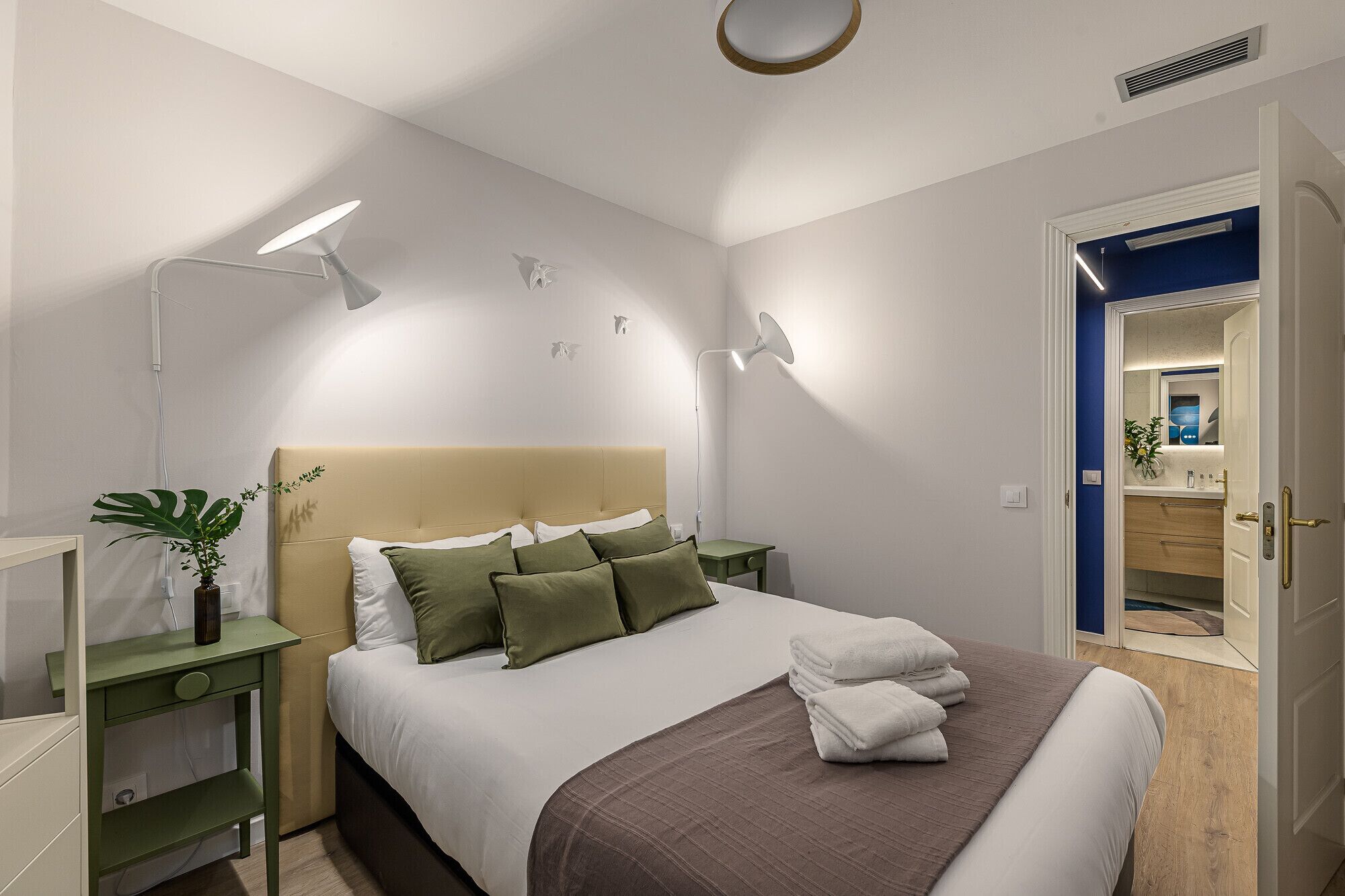 A modern bedroom with a large bed, bedside tables, a nightstand, a potted plant, and a bathroom visible through an open door.