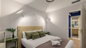 A modern bedroom with a large bed, bedside tables, a nightstand, a potted plant, and a bathroom visible through an open door. - Waou La Latina V - Short Term Rental (Madrid)