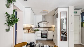A compact kitchen with white cabinets, a built-in oven, and a dining area with two chairs and a table set for two. - Waou La Latina V - Short Term Rental (Madrid)