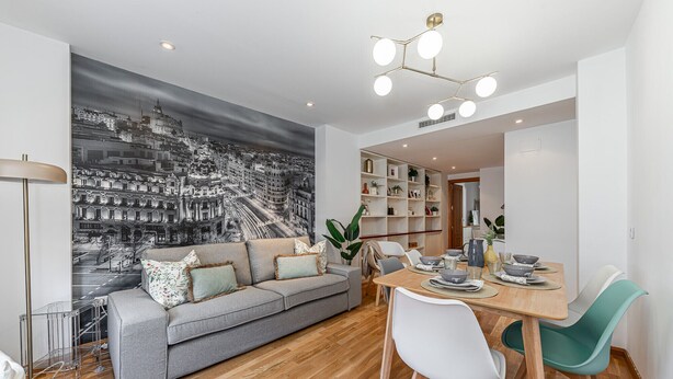 A modern living room with a grey sofa, wooden dining table, and teal chairs. A cityscape mural adorns the wall, and a bookshelf is visible in the background. - Waou Chamberi X - Short Term Rental (Madrid)