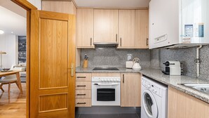 A compact kitchen with wooden cabinets, a built-in oven, a washing machine, and a countertop with a kettle and toaster. - Waou Chamberi X - Short Term Rental (Madrid)