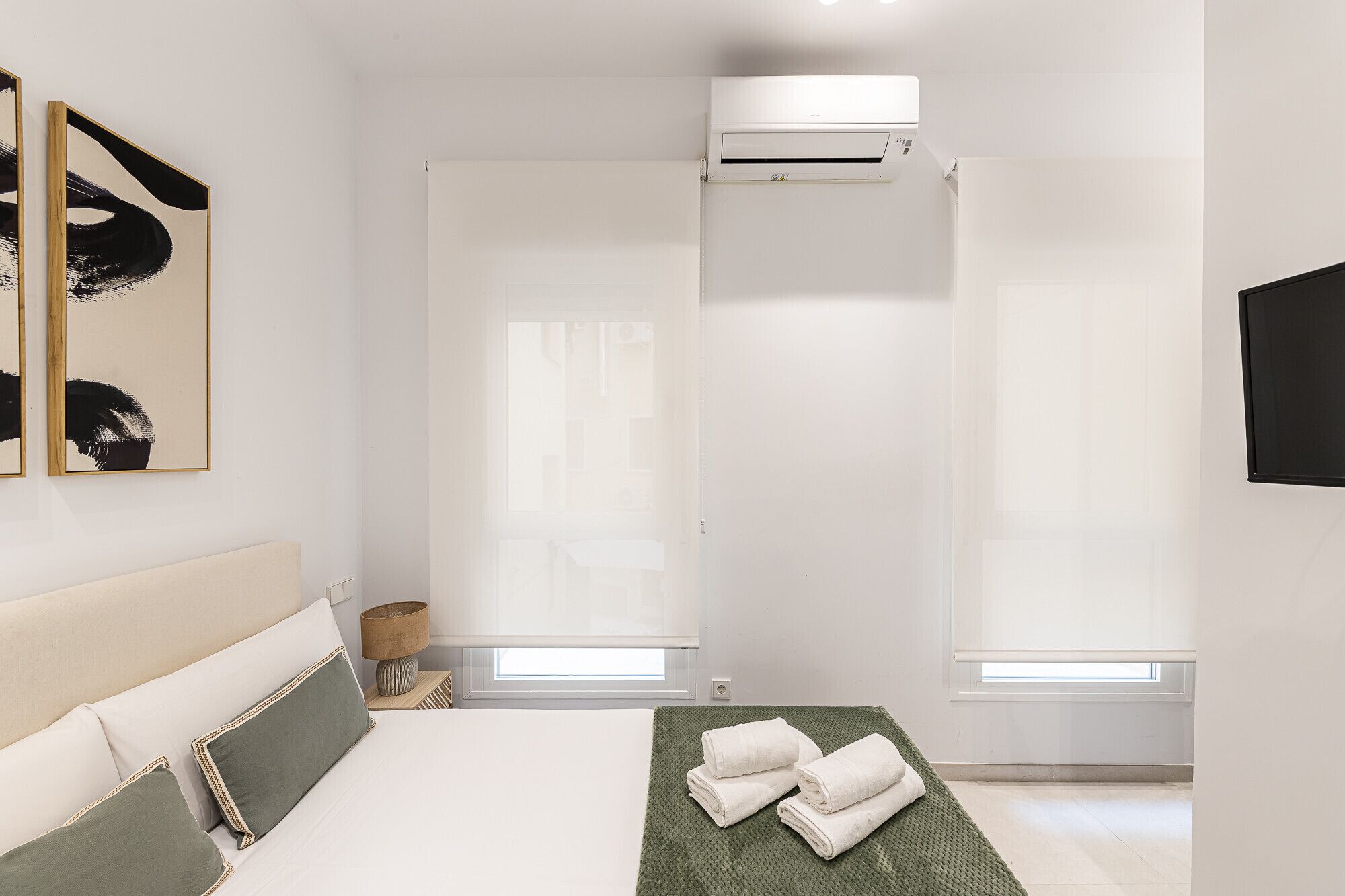 A modern bedroom with a bed, two windows, a wall-mounted air conditioner, and artwork on the wall.