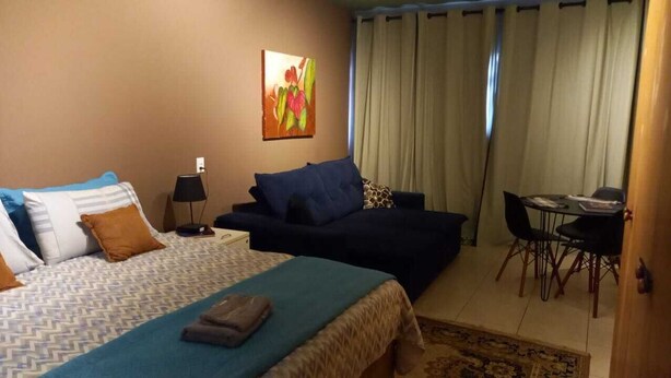A hotel room with a bed, a sofa, a desk with chairs, and a painting on the wall. - Studio Barreto Leme 72 (Campinas)