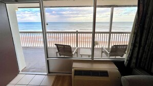 A room with a large window overlooking a beach. There are two chairs and a small table on the balcony. - Beachfront Studio - King Bed & Direct Ocean View (Daytona Beach)