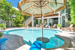 A swimming pool with a thatched umbrella, lounge chairs, and a staircase leading to an upper level.