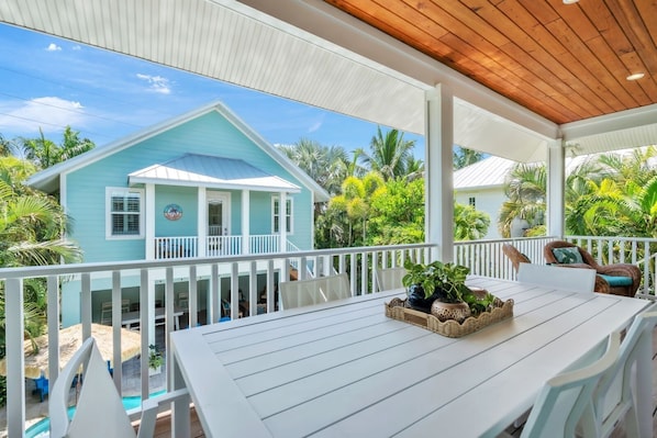 A covered outdoor area with a table and chairs, a view of a turquoise house, and surrounding tropical vegetation.