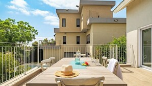 Una terraza con una mesa lista para dos personas, una piscina al fondo y un edificio de varios pisos.