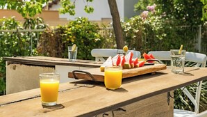 Un área de comedor al aire libre con una mesa de madera, dos vasos de jugo de naranja, una sandía y un limón sobre una tabla de madera, y un vaso de agua.