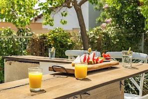 Area pranzo all'aperto con tavolo in legno, due bicchieri di succo d'arancia, un'anguria e un limone su un tagliere, e un bicchiere d'acqua.
