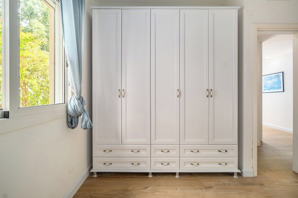 A white wardrobe with multiple drawers and doors, positioned next to a window with a curtain.