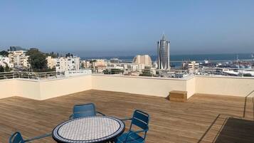 Una terraza en la azotea con una mesa redonda y sillas, con vistas a un paisaje urbano que incluye edificios y el mar.